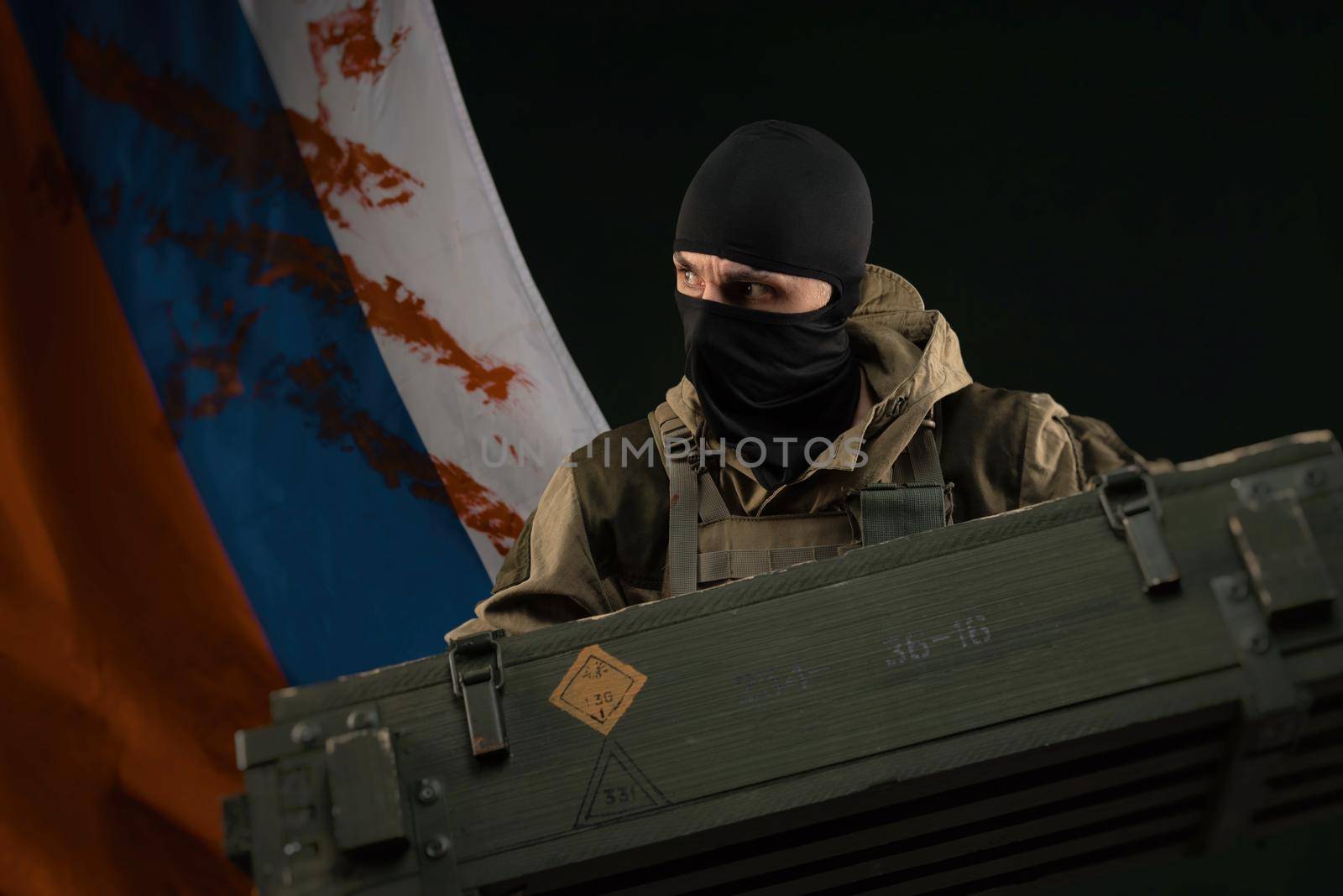 male soldier in a military uniform with a balaclava holds a box of ammunition and grenades against the background of the bloody flag of the Russian Federation