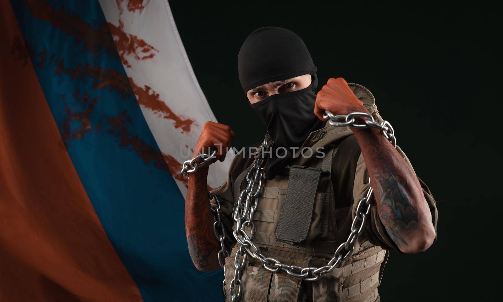 a male soldier in a military uniform with a balaclava and hands covered in blood holds a chain as a symbol of slavery against the background of the bloody flag of Russia by Rotozey