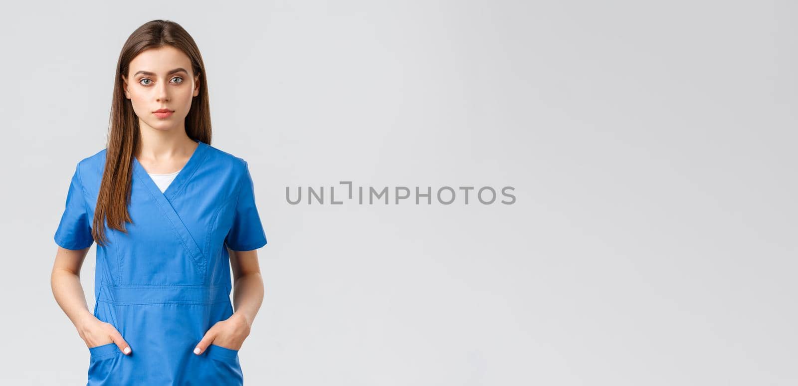 Healthcare workers, prevent virus, insurance and medicine concept. Serious-looking, confident young medical worker, nurse or doctor in blue scrubs hold hands in pockets and looking camera.