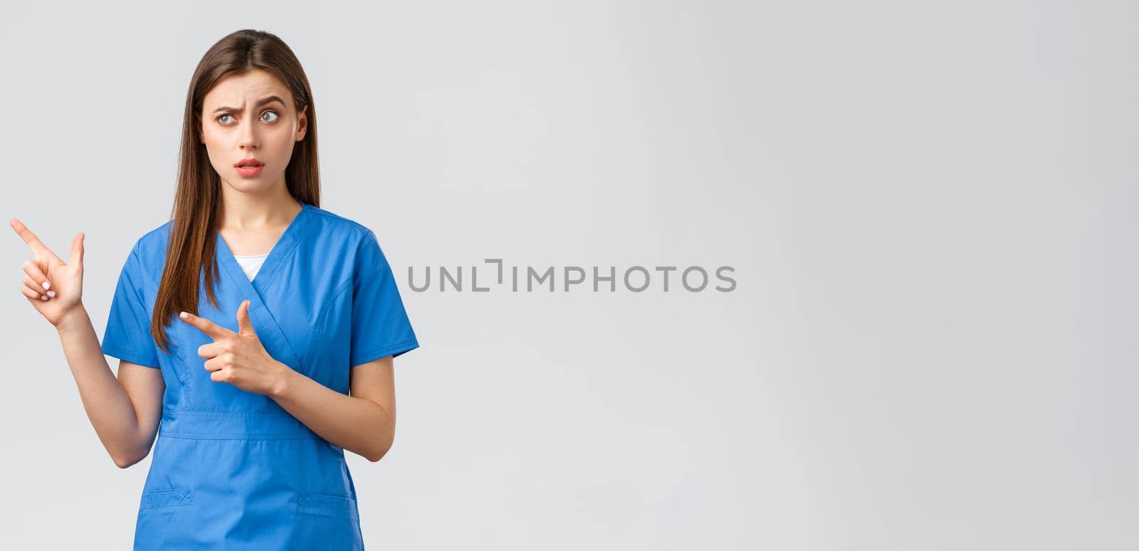 Healthcare workers, prevent virus, insurance and medicine concept. Skeptical and unsure nurse or doctor in blue scrubs, raise eyebrow suspicious, pointing fingers left at banner by Benzoix