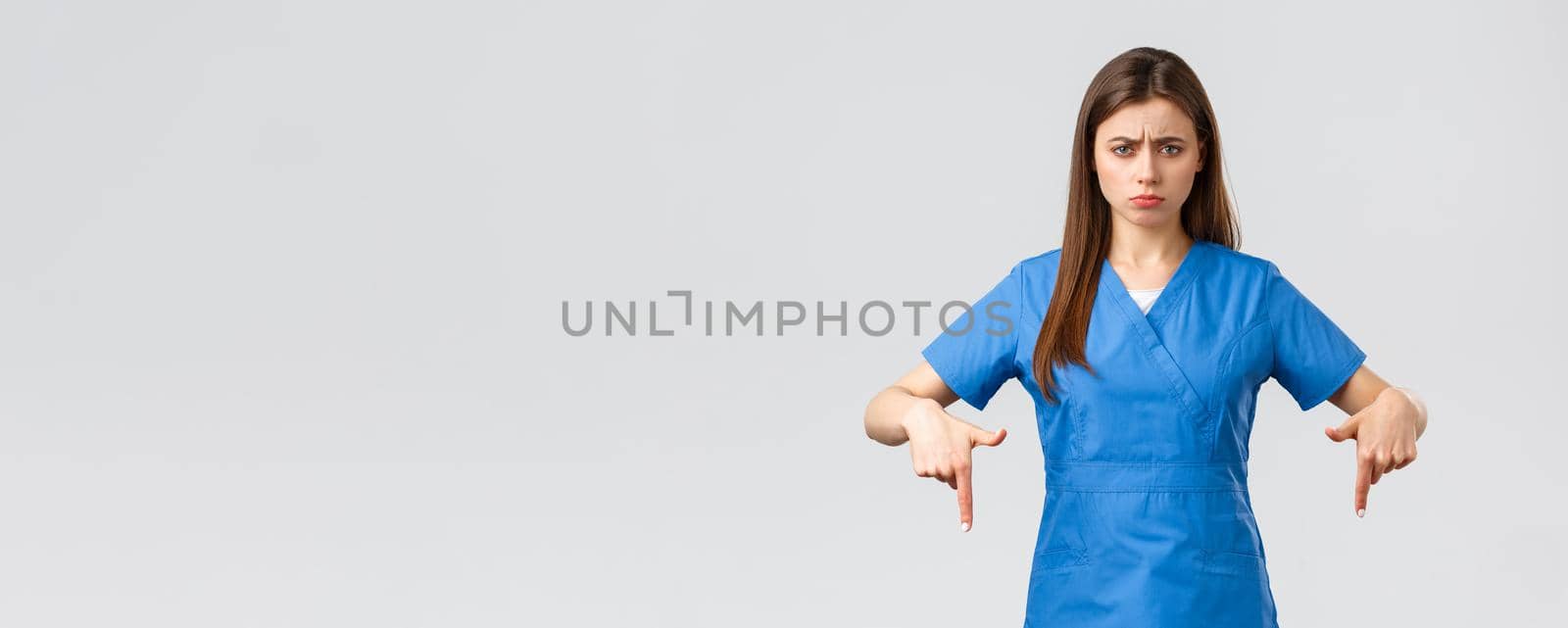 Healthcare workers, prevent virus, covid-19 test screening, medicine concept. Disappointed and uneasy cute nurse or doctor in blue scrubs, pointing fingers down, showing bad unpleasant news by Benzoix