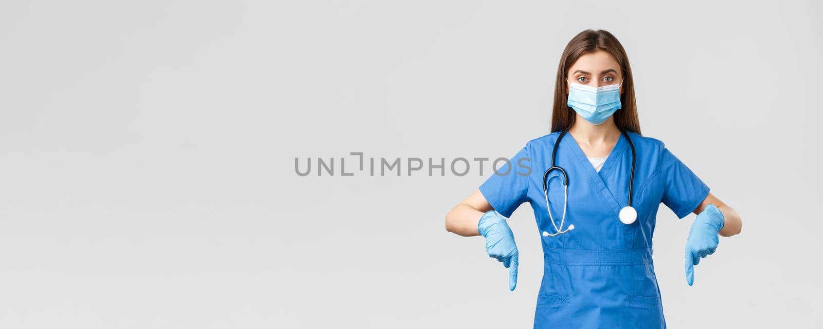 Covid-19, preventing virus, health, healthcare workers and quarantine concept. Determined female nurse or doctor in blue scrubs, medical mask and gloves, pointing fingers down, showing info banner.