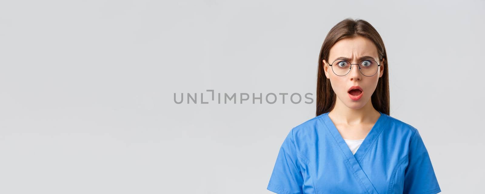 Healthcare workers, medicine, insurance and covid-19 pandemic concept. Shocked concerned female nurse, doctor in blue scrubs and glasses, say oh my gosh frustrated, open mouth and frowning by Benzoix