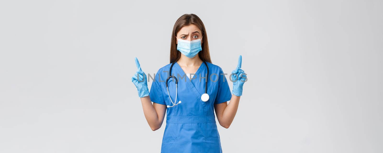 Covid-19, preventing virus, health, healthcare workers and quarantine concept. Skeptical female nurse in blue scrubs and medical mask dont believe this info, pointing fingers up doubtful.