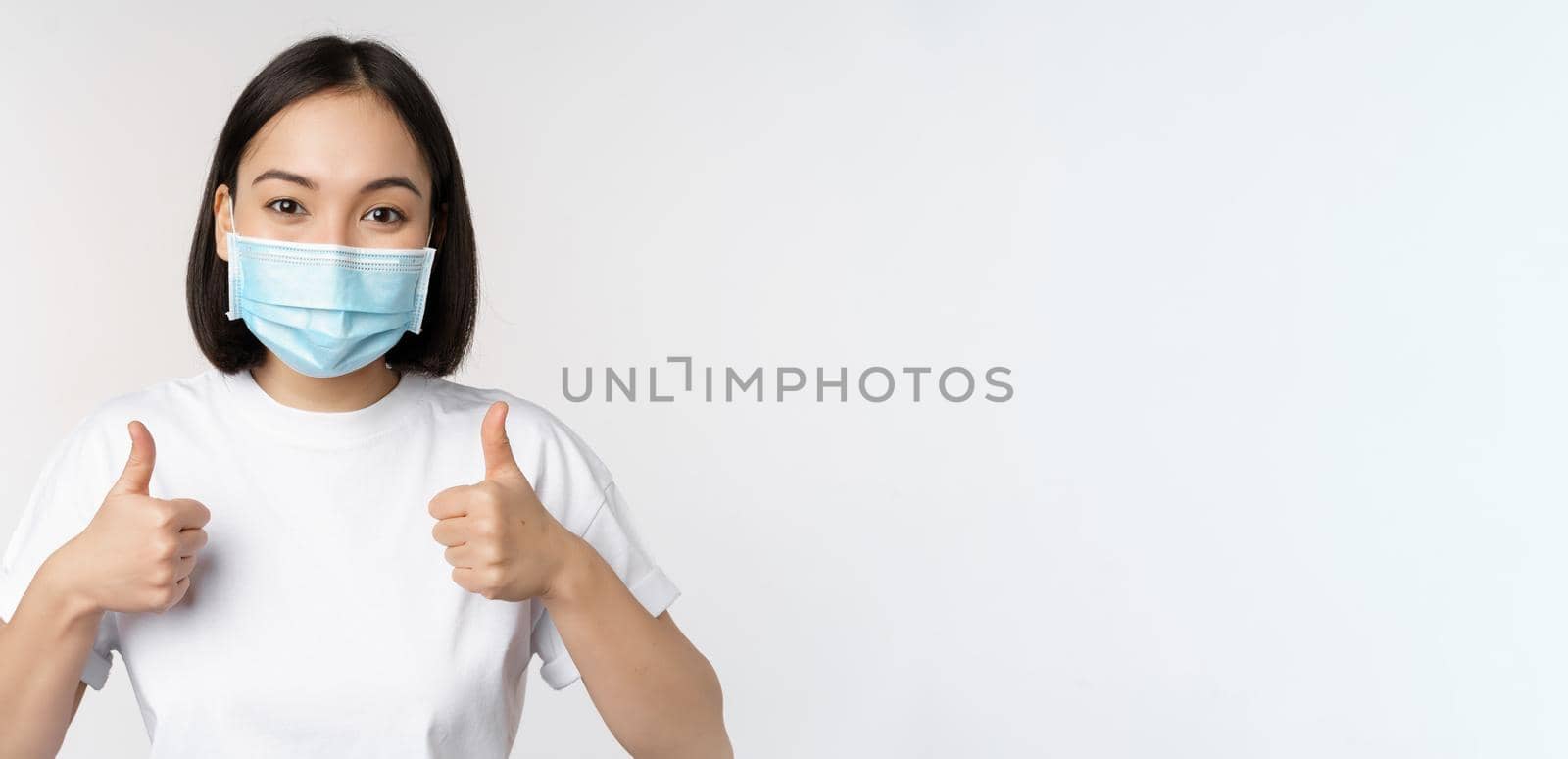 Covid-19, health and people concept. Young happy korean woman in medical mask, showing thumbs up, standing in tshirt over white background by Benzoix