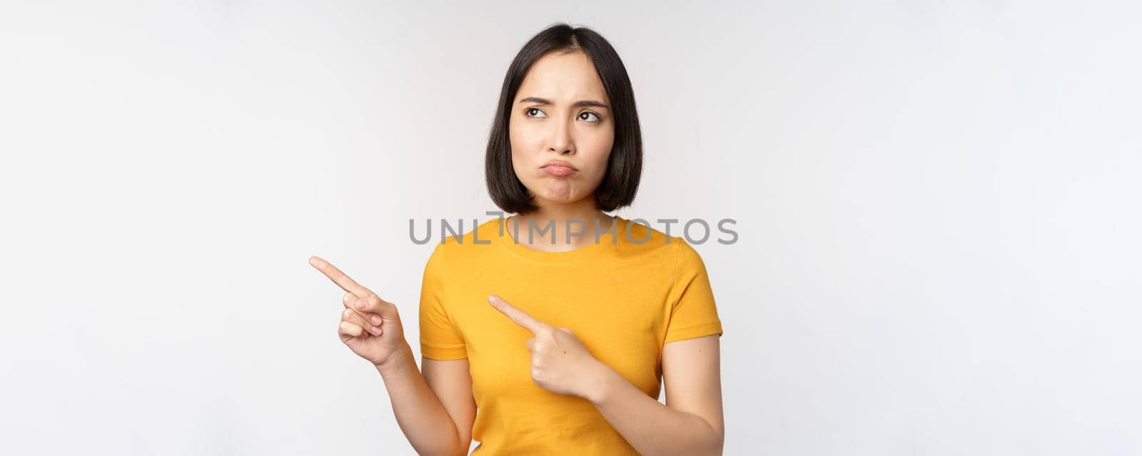 Upset grumpy asian girl, pouting and looking at smth unfair, pointing fingers left at banner, logo brand, standing in yellow tshirt over white background.