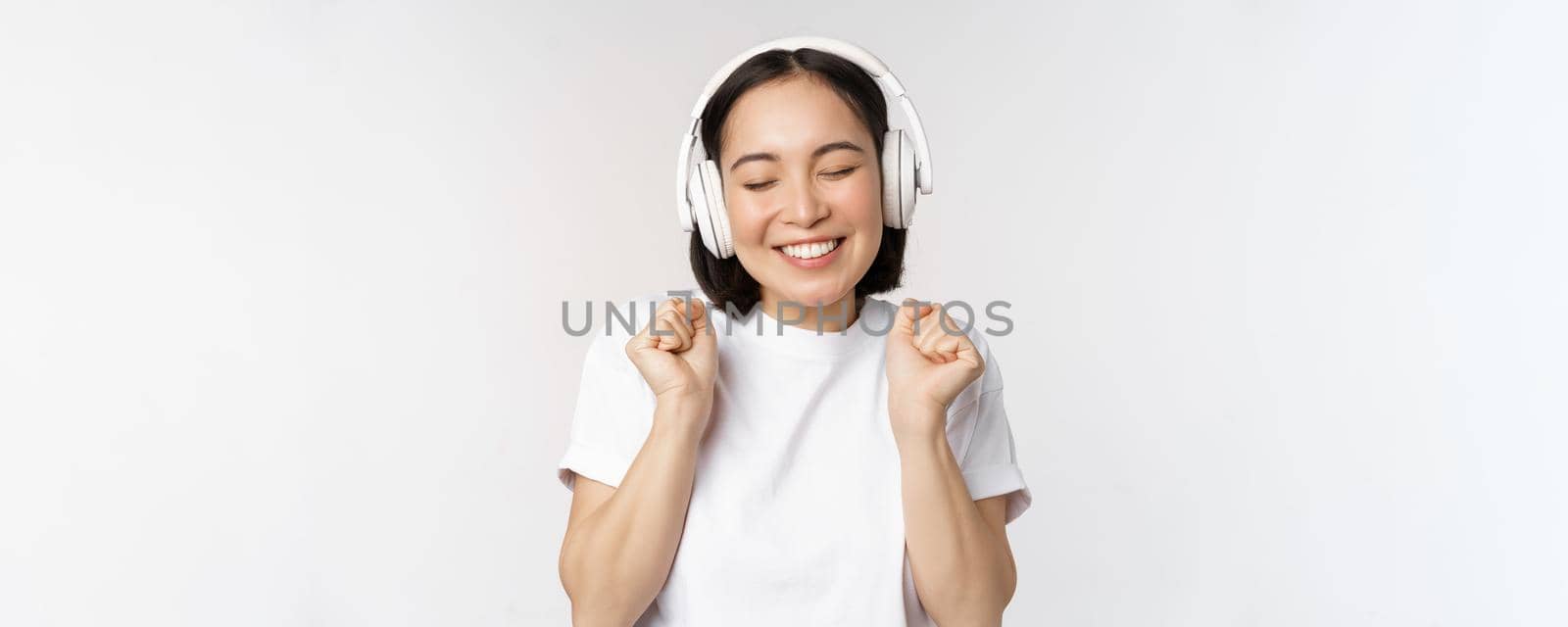 Modern asian girl dancing, listening music with headphones, smiling happy, standing in tshirt over white background by Benzoix