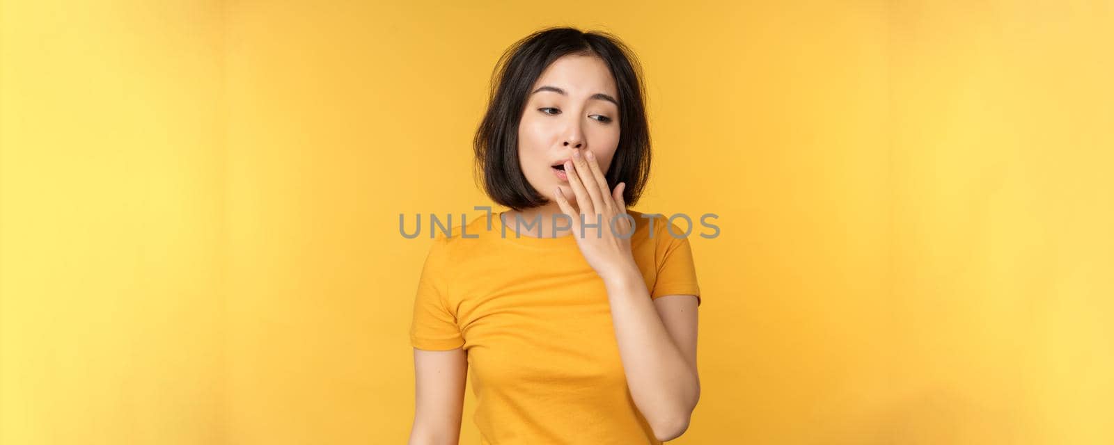 Image of young asian girl student, yawning, feeling tired, standing sleepy agaisnt yellow background by Benzoix