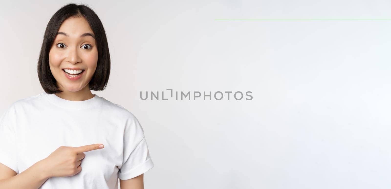 Happy beautiful asian girl showing advertisement on empty copy space, pointing finger right and smiling, demonstrating banner or company logo, white background.