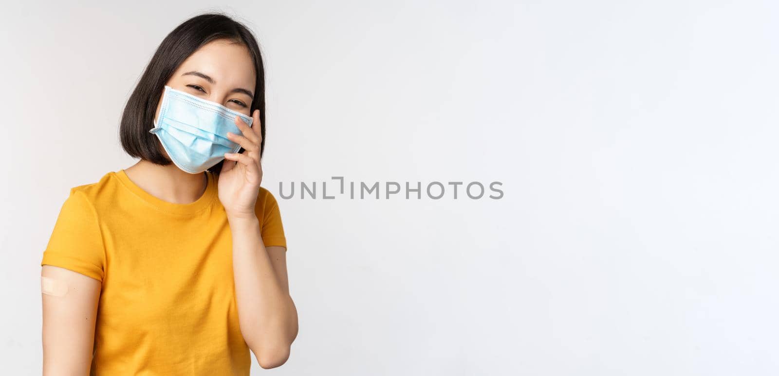 Covid-19, vaccination and healthcare concept. Cute asian girl in medical face mask, showing band aid after coronavirus vaccination, standing over white background by Benzoix