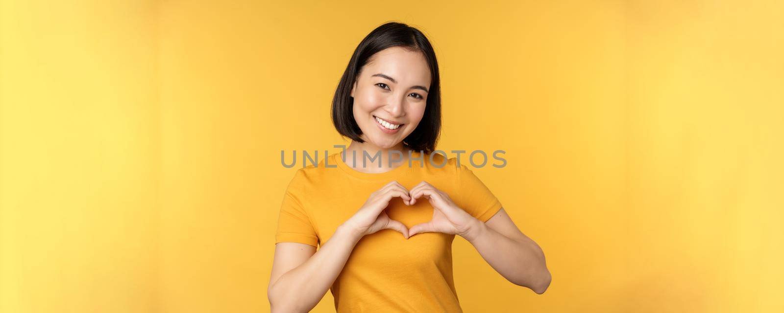 Beautiful asian girl showing heart, love gesture and smiling white teeth, express care and sympathy, standing over yellow background.