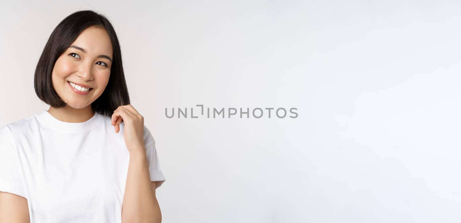 Portrait of cute coquettish woman laughing and smiling, looking aside thoughtful, thinking or imaging smth, standing in white t-shirt over studio background.