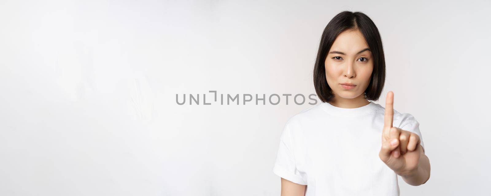 Image of asian girl showing stop, prohibit smth, extend one arm to show forbidding, taboo gesture, standing in tshirt over white background.