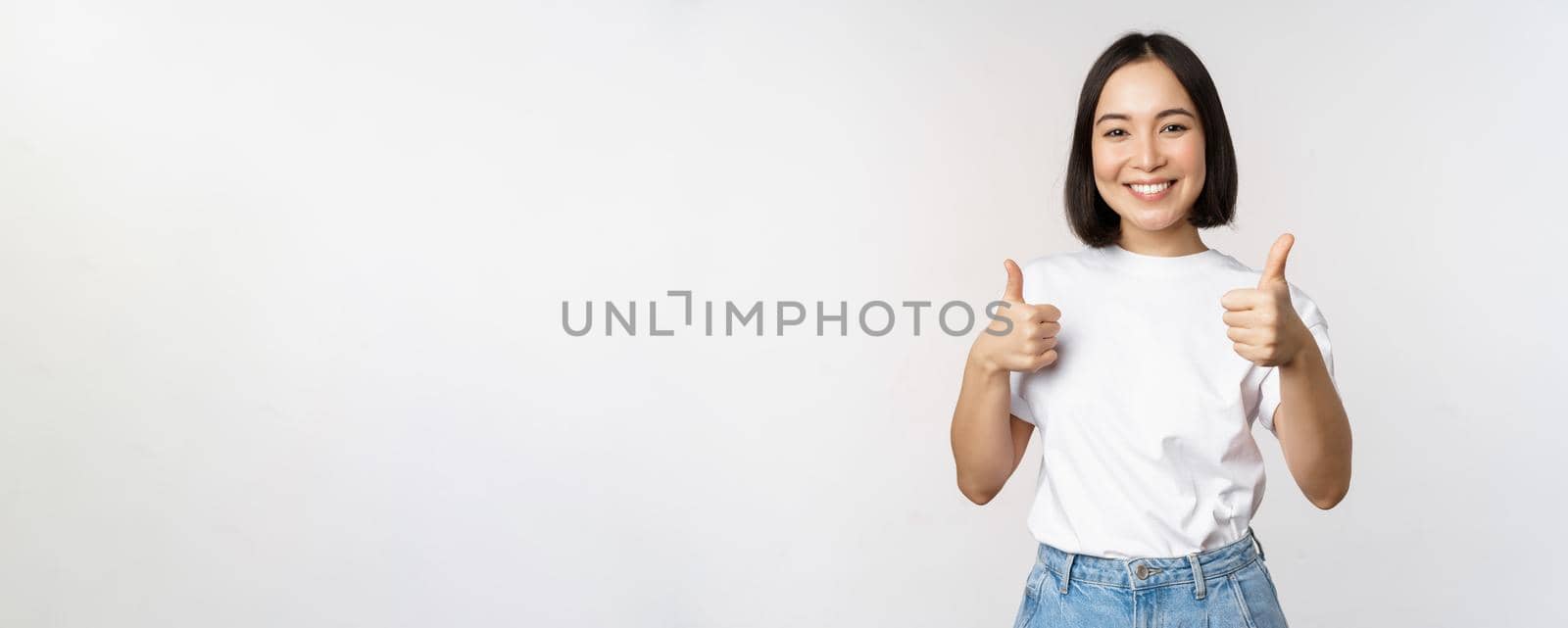 Happy beautiful korean girl, showing thumbs up in approval, smiling pleased, like smth, recommending, standing over white background.