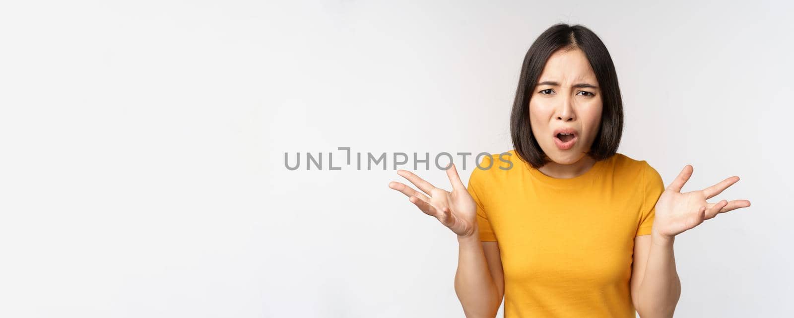 Frustrated, confused asian woman cant understand smth, looking clueless, shrugging shoulders, standing in yellow tshirt over white background by Benzoix