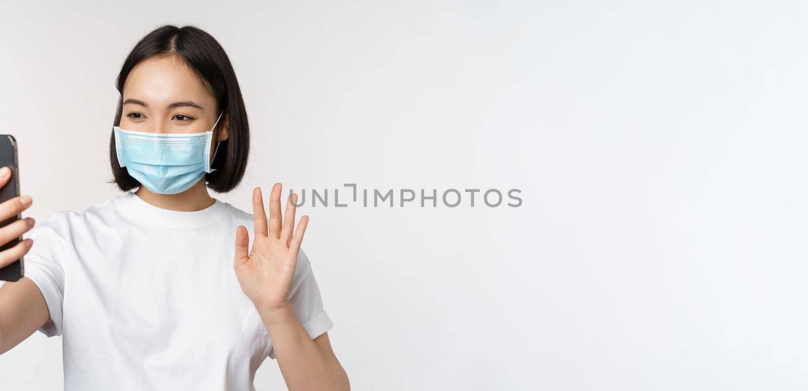 Health and covid-19 concept. Modern asian girl, student in medical mask, video chat with mobile phone, waving hand at smartphone app, standing over white background.