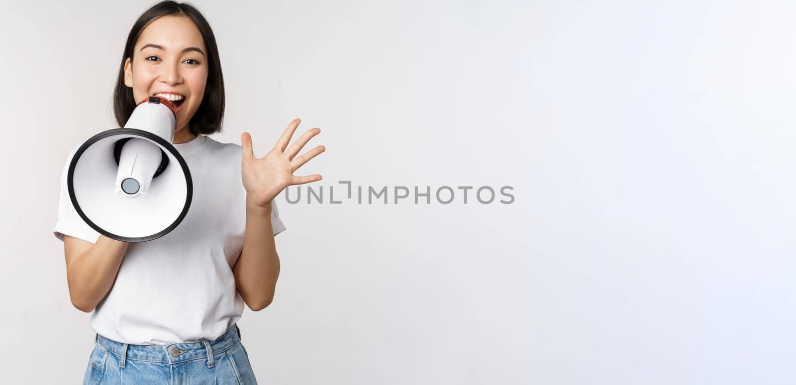 Happy asian woman shouting at megaphone, making announcement, advertising something, standing over white background.