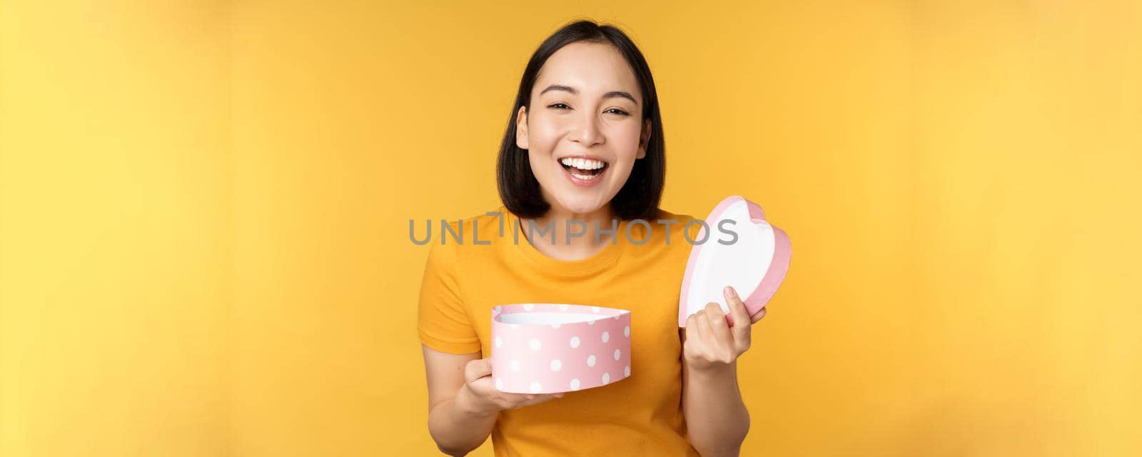 Portrait of excited asian woman, open gift box with surprised happy face, standing over yellow background.