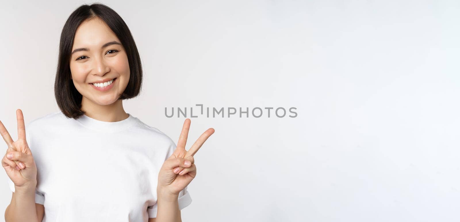 Cute asian girl showing peace, v-sign, smiling and looking happy at camera, wearing white tshirt, studio background by Benzoix