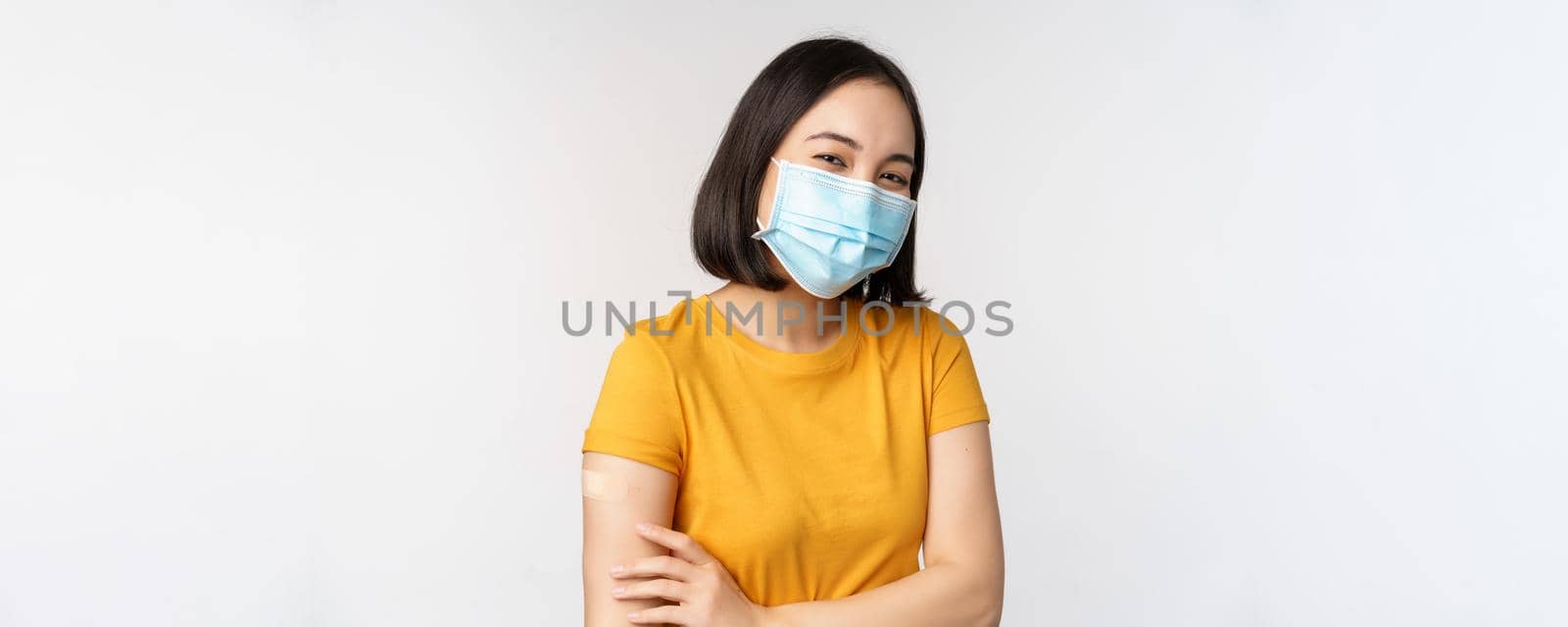 Covid-19, vaccination and healthcare concept. Cute asian girl in medical face mask, holding shoulder with band aid, got vaccine from coronavirus, smiling pleased, white background.
