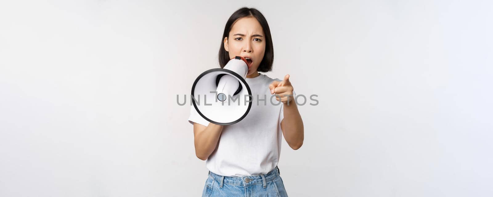 Angry asian woman with megaphone, scolding, accusing someone, protesting with speakerphone on protest, standing over white background.
