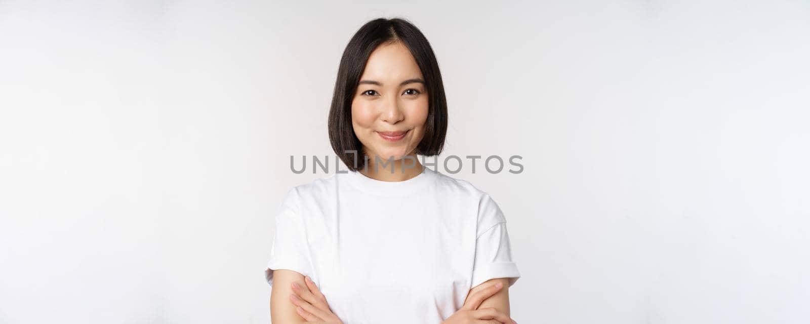 Close up portrait of confident korean girl, student looking at camera with pleased smile, arms crossed on chest, standing over white background by Benzoix