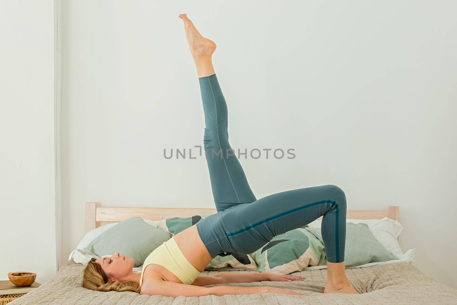 A slender woman in green leggings does yoga in the morning, lying on her back, lifting her leg up, at home, on the bed. copy space