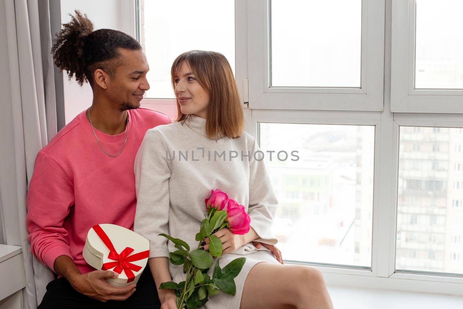 Happy enamored guy in a pink jumper and a girl in a white dress sit by the window, hold a white heart-shaped box with a bow and a bouquet of roses. copy space