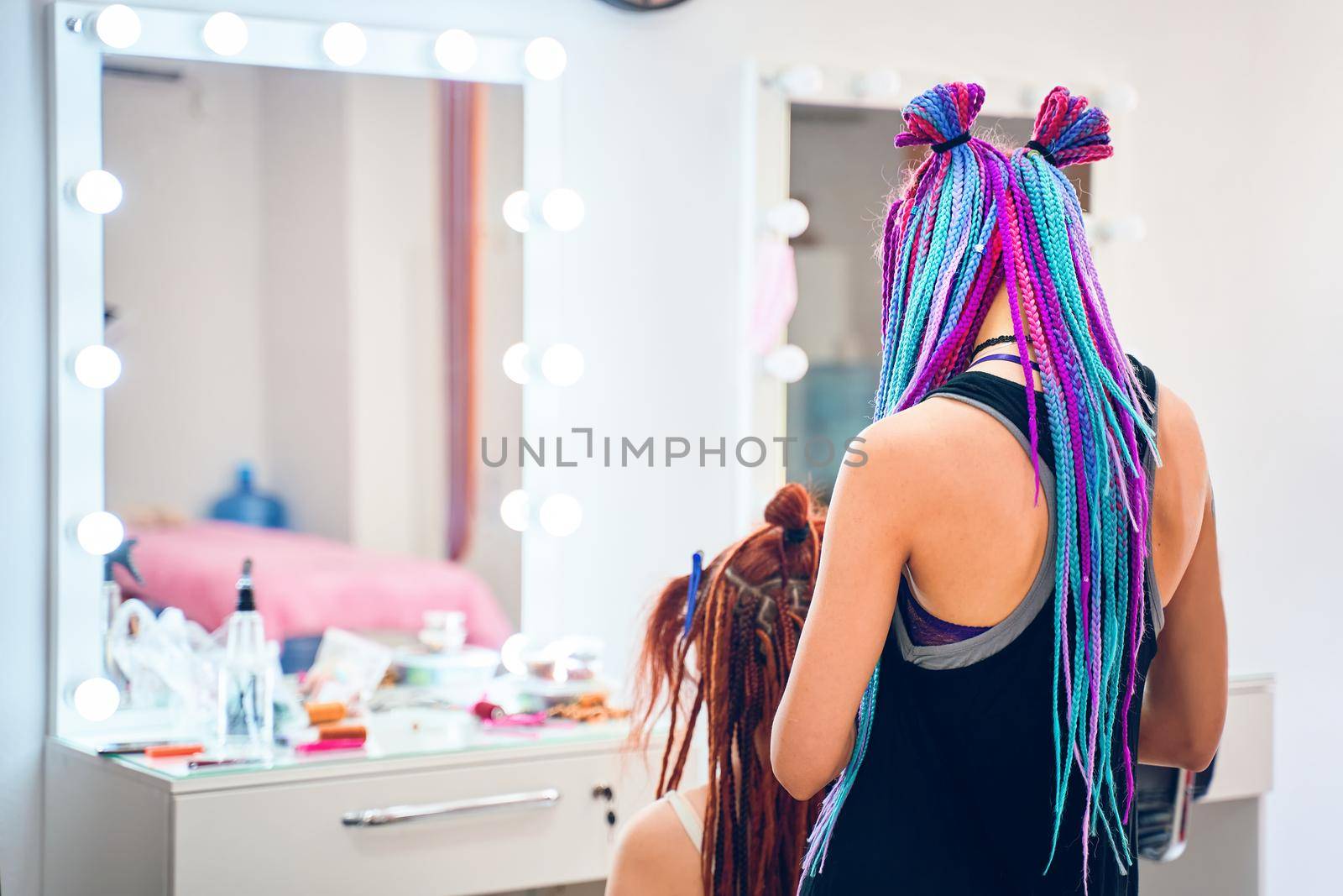 Back view of female barber with colored pigtails, who braids ginger dreadlocks to woman. Creative and fashionable hairstyle. Service in beauty salon. Large mirror with light bulbs on background.