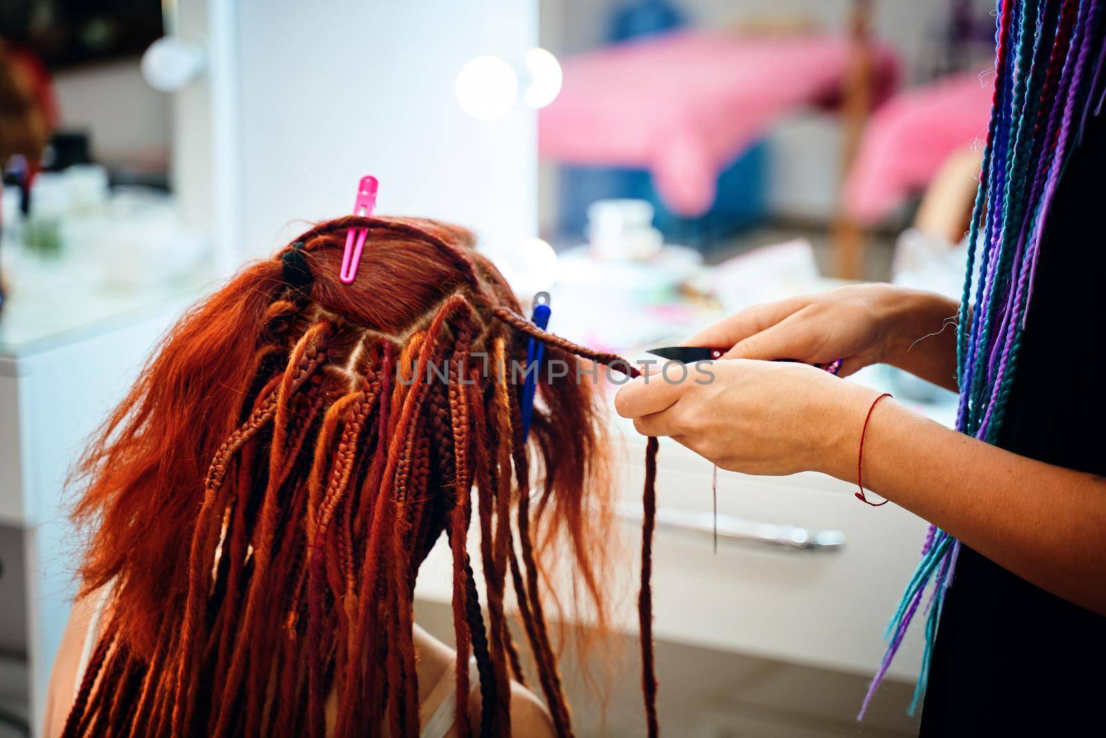 Braiding process african plaits with colored kanekalon. Girl hairdresser weaves woman ginger dreadlocks. Bright pigtails on head. Hippie style hairstyle. Beauty salon services.