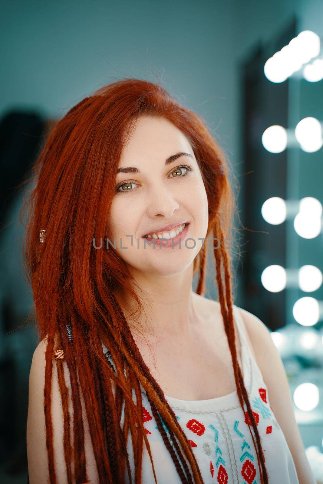 Smiling red-haired girl with long dreadlocks. Beautiful woman looks at camera. Portrait of female with ginger dreads in beauty salon. Unusual coiffure. Hippie or boho style.