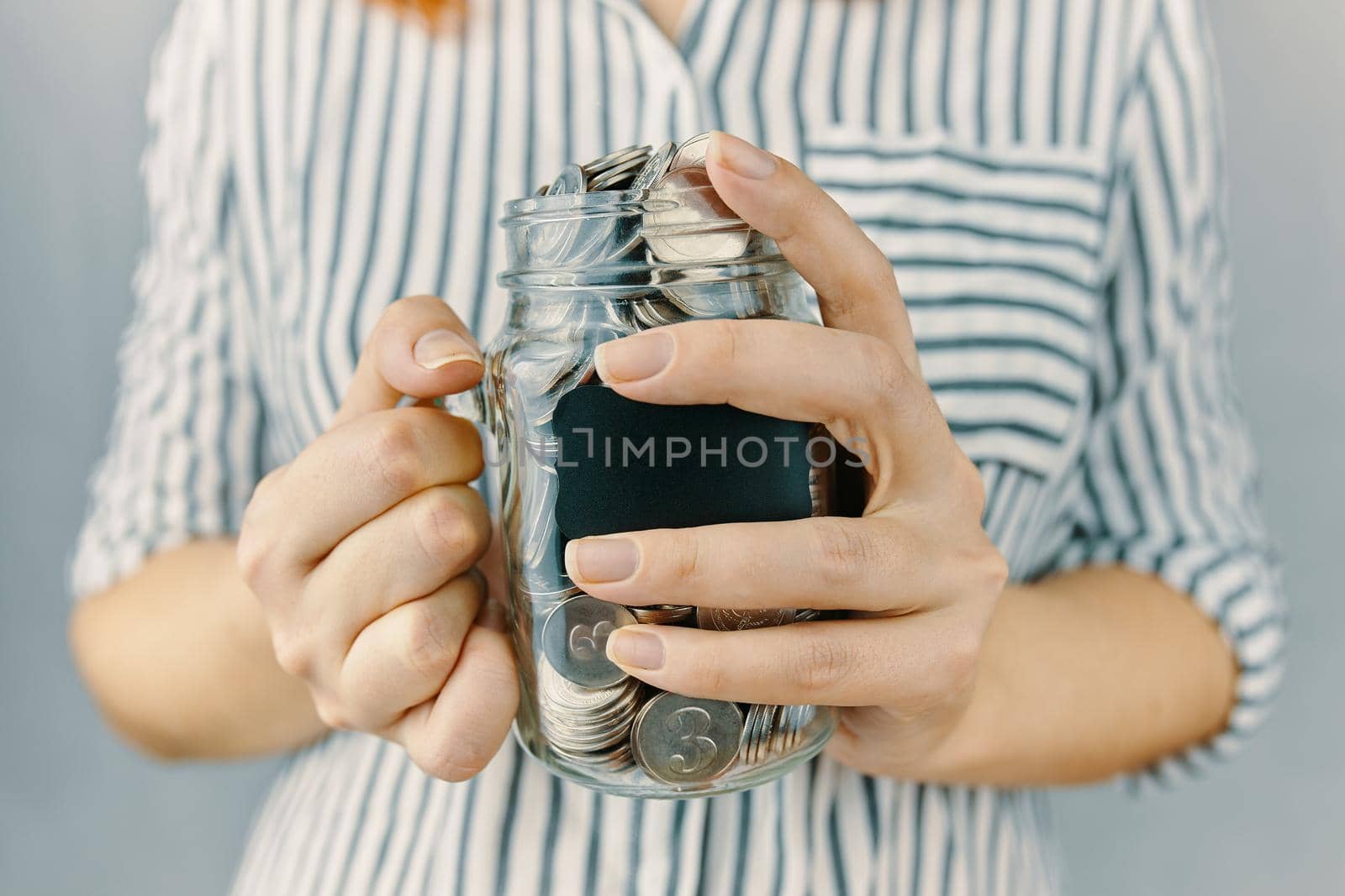 Glass bottle with coins in women's hands. Blank black sticker on donation money jar. Girl in white and black striped shirt holds her savings. Pot for tips.