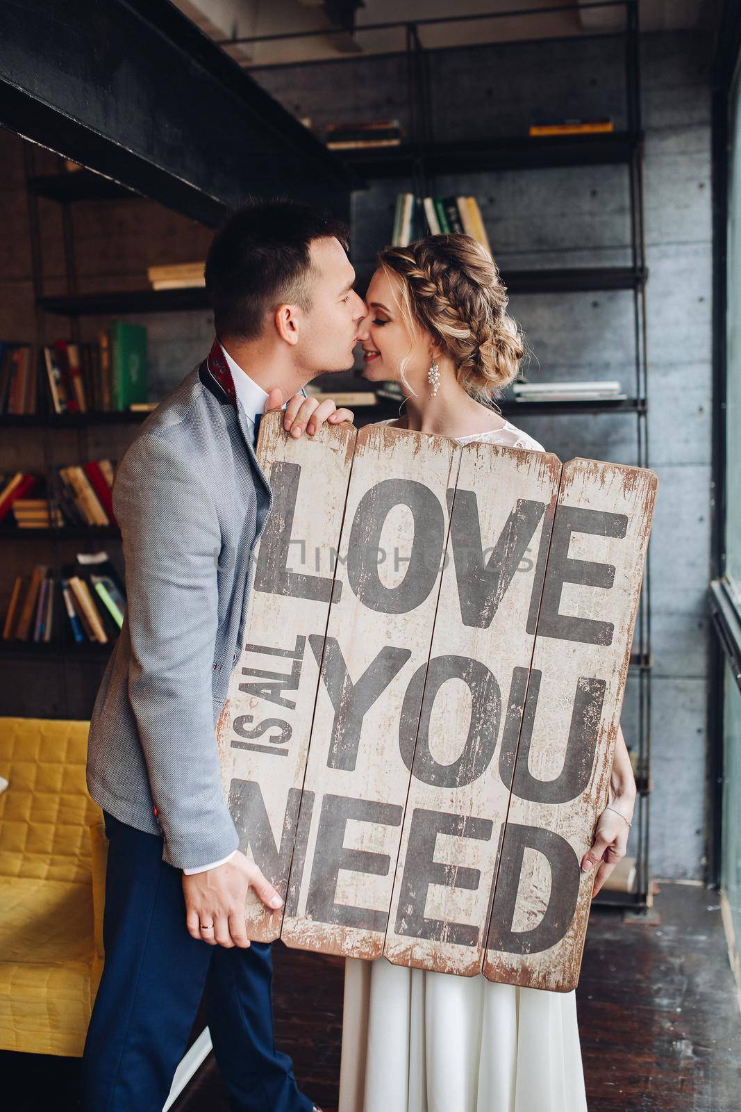 Portrait of lovely newlywed couple holding wooden signboard with message all you need is love. Husband kissing his wife in bookshop.