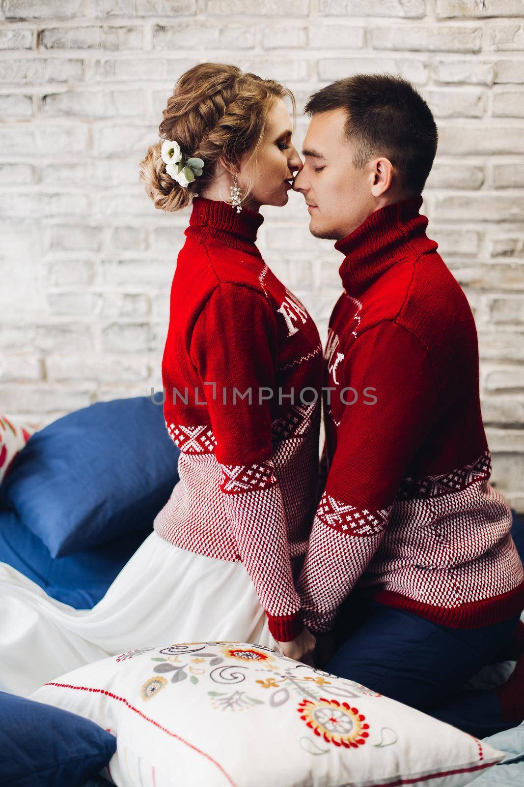 Couple in red Christmas jumpers kissing surrounded by pillows. by StudioLucky