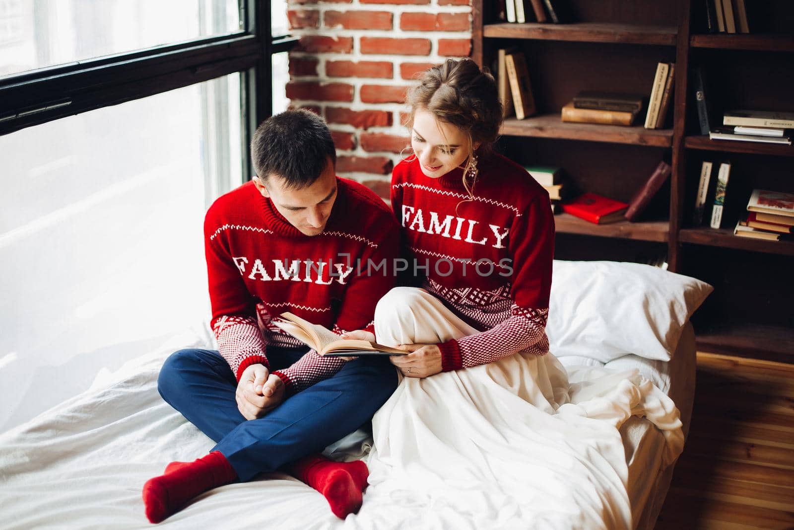 View of young couple in red sweaters reading book together near big window, smiling, spanding time together. Boyfriend with girlfriend sitting in bed. Family celebrating new year together. Students.