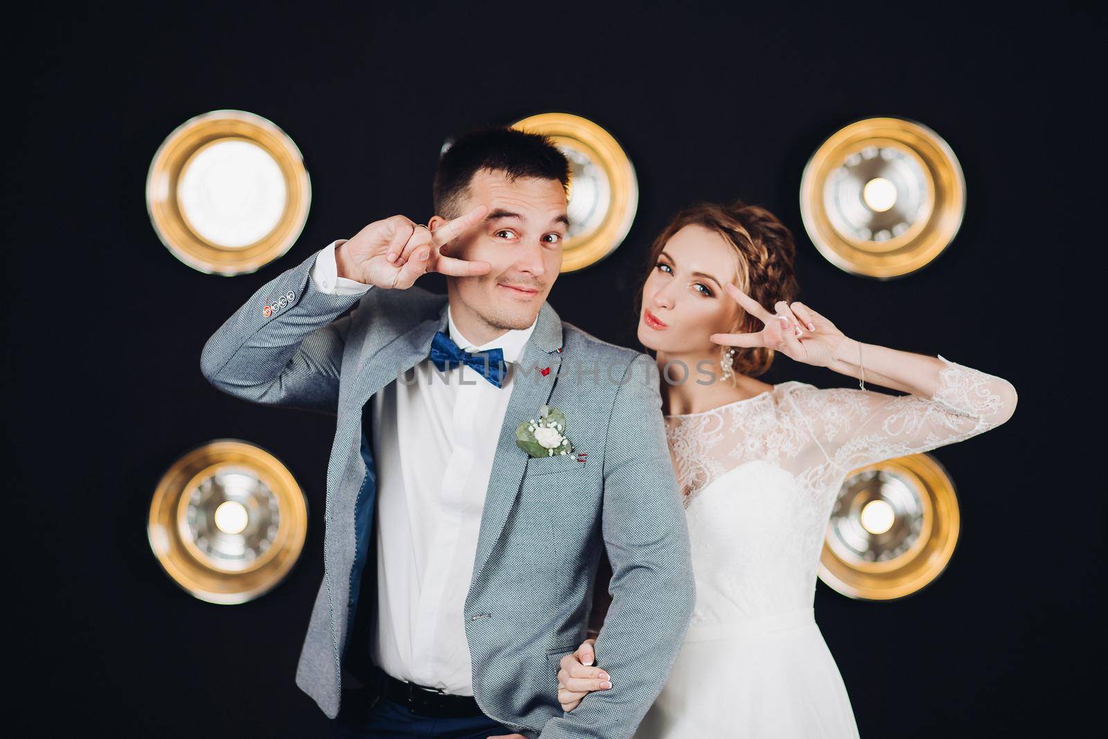 Studio portrait of two funny and positive wedding couple of groom and bride having fun, dancing at party, showing peace by fingers. Beautiful young married couple having fun together.
