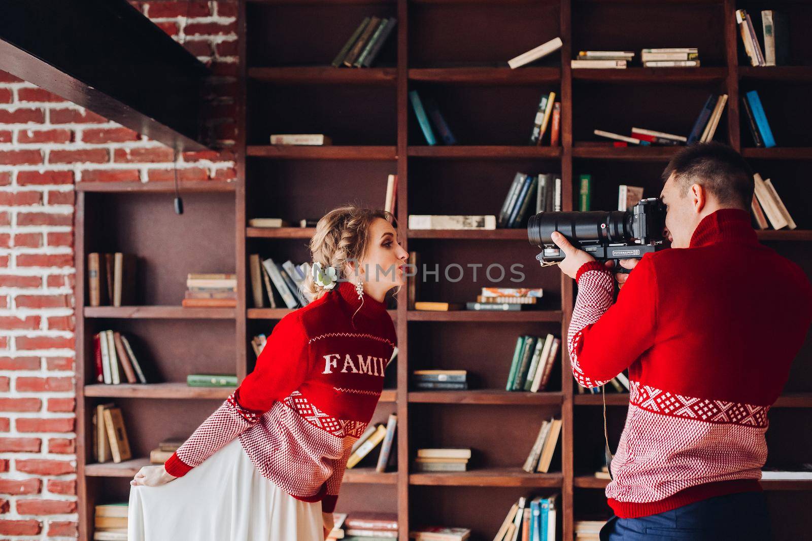 Side view of newlywed in similar red sweater with family word having fun at home. Wife posing to her husband taking photos of her with camera.