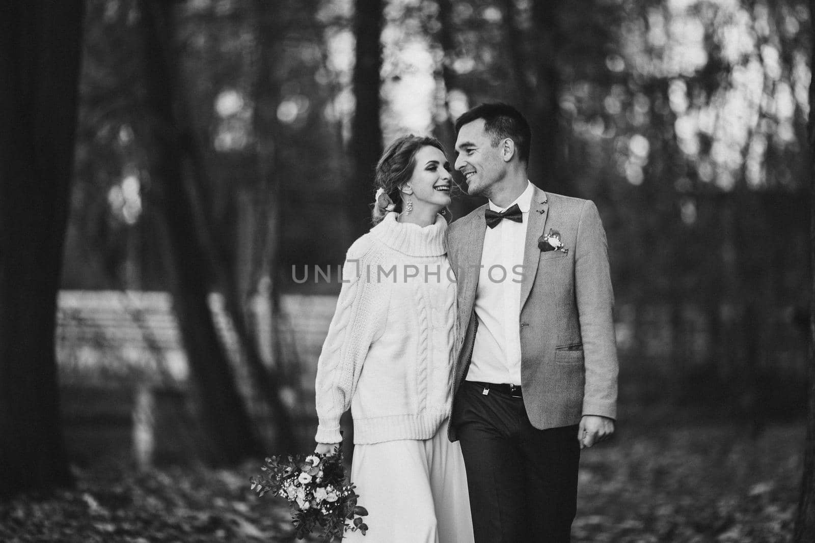 Black and white portrait of cheerful beautiful newlywed couple walking in forest smiling. Bride wearing cute cozy sweater and holding bridal bouquet. Bokeh.