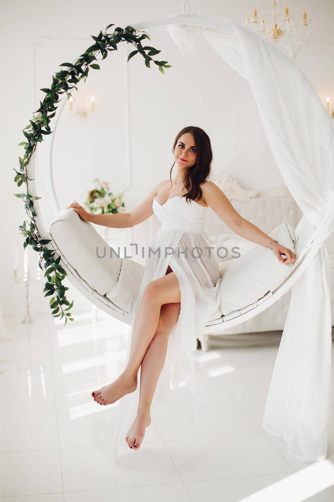 Full length portrait of stunning pregnant brunette woman in elegant white dress with transparent skirt sitting on modern suspended chair in beautifully decorated white bedroom. She is looking at camera.