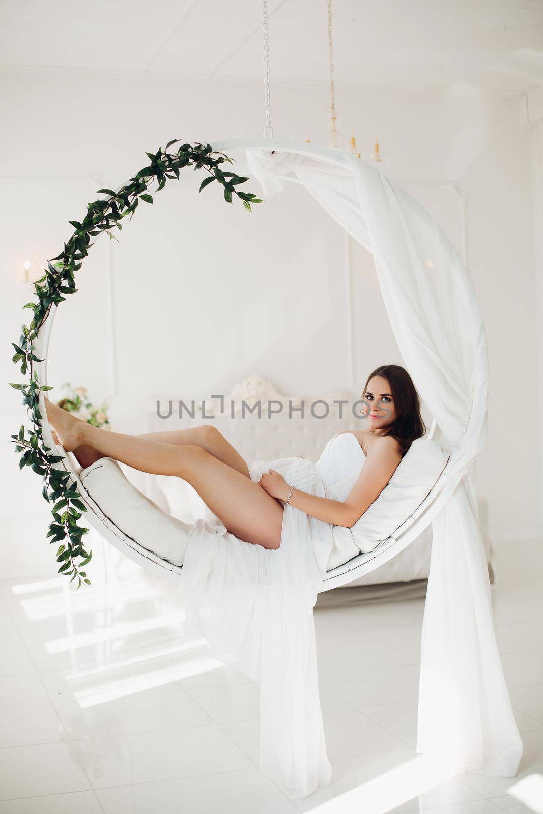 Full length portrait of stunning pregnant brunette woman in elegant white dress with transparent skirt sitting on modern suspended chair in beautifully decorated white bedroom. She is looking at camera.