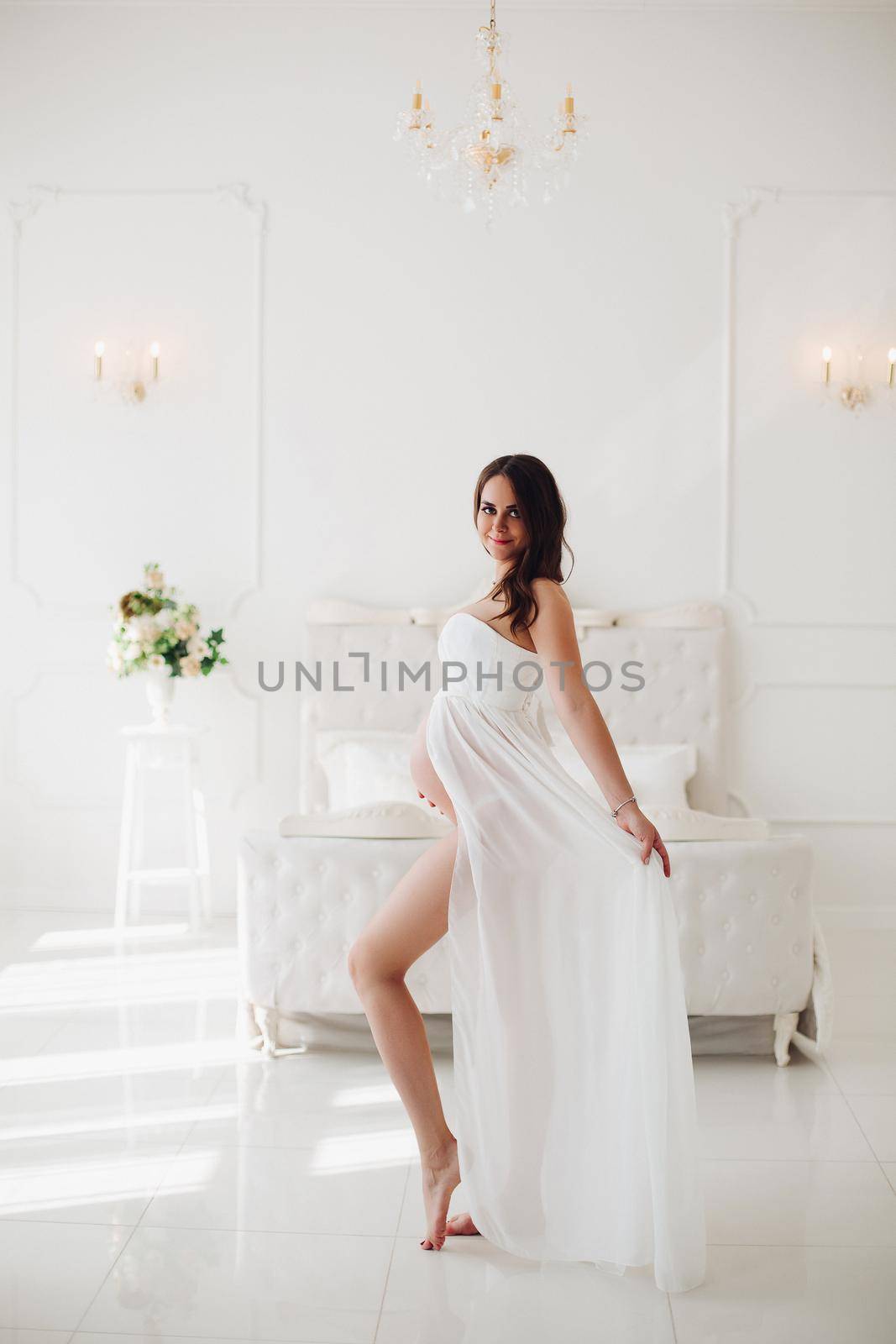 View from side of future mother in beautiful white dress standing in bedroom, looking at camera, smiling and posing. Adorable pregnant woman expecting little child. Concept of pregnancy and love.