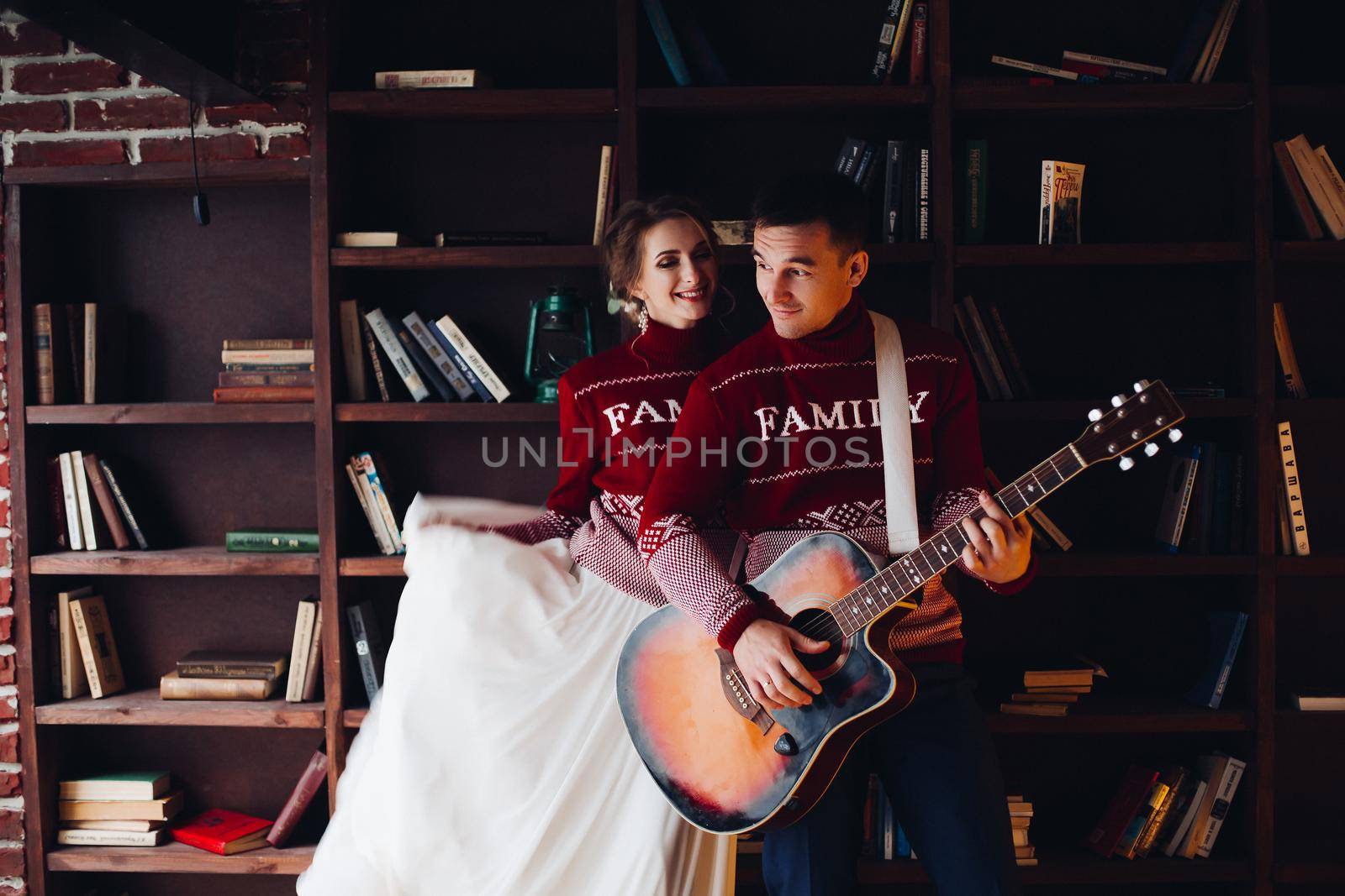 Portrait of husband and wife posing happily in red sweater with family word. Man holding guitar and woman flattering her wedding dress skirt.