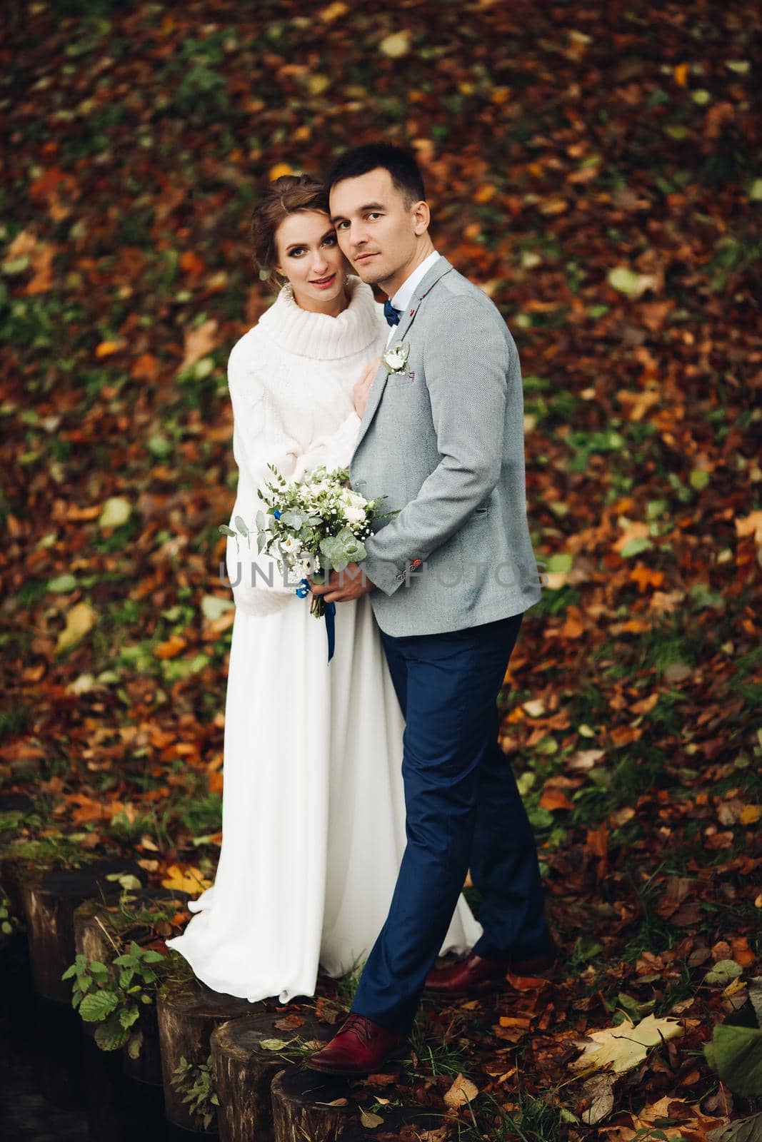 Back view of husband embracing his beautiful elegant wife in lace wedding dress with lovely bouquet against unfocused lake surface.