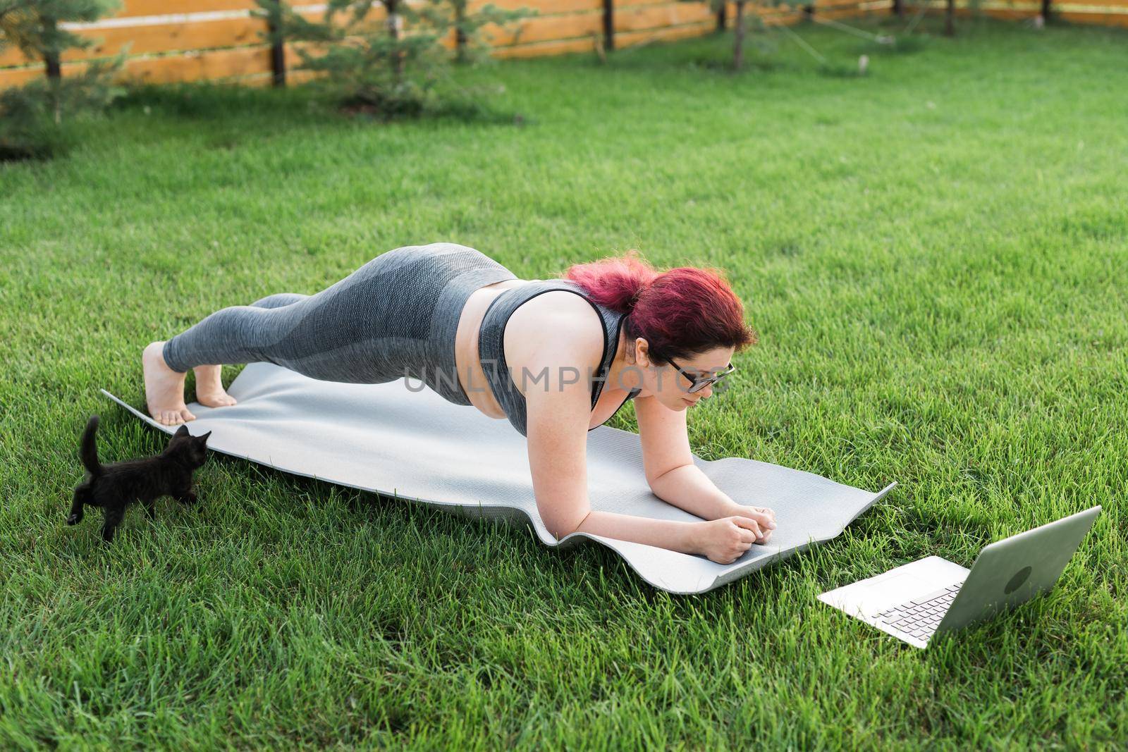 Young plus size woman in sporty top and leggings standing in plank on yoga mat spending time on green grass in yard. Black kitten walks around her. Well being fitness concept