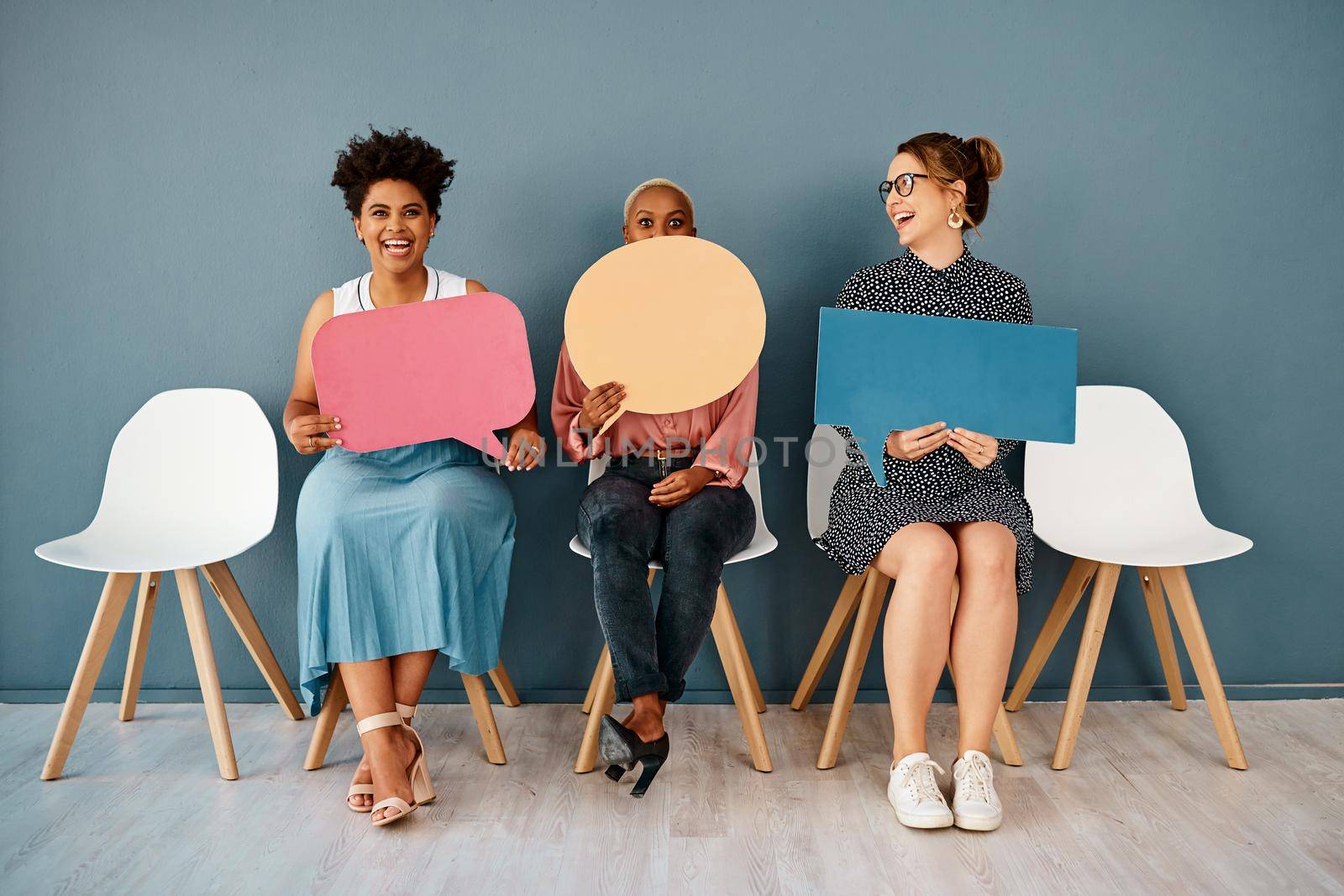 Studio shot of a group of attractive young businesswomen holding speech bubbles while sitting in a row against a grey background.