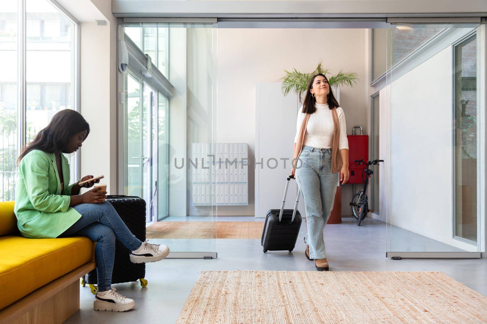 Happy young caucasian woman enters hotel reception with trolley suitcase. Copy space. by Hoverstock
