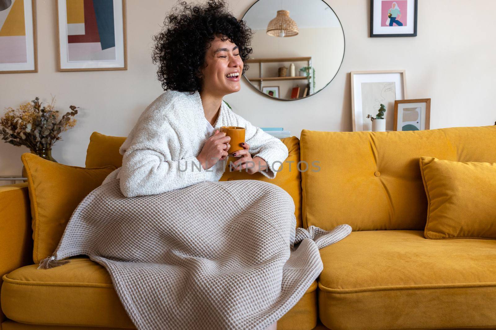 Happy African american woman enjoying quiet time at home laughing, drinking morning coffee sitting on sofa. Copy space. by Hoverstock