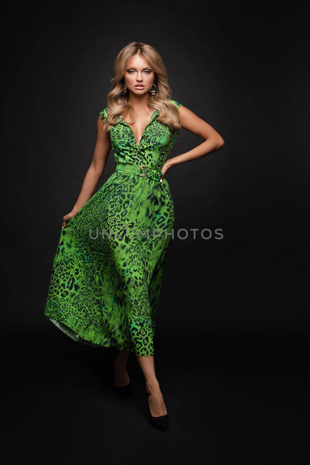 Front view of slim blonde standing on black isolated background and posing. Attractive young woman in green long dress looking at camera and smiling in studio. Concept of elegance and glamour.