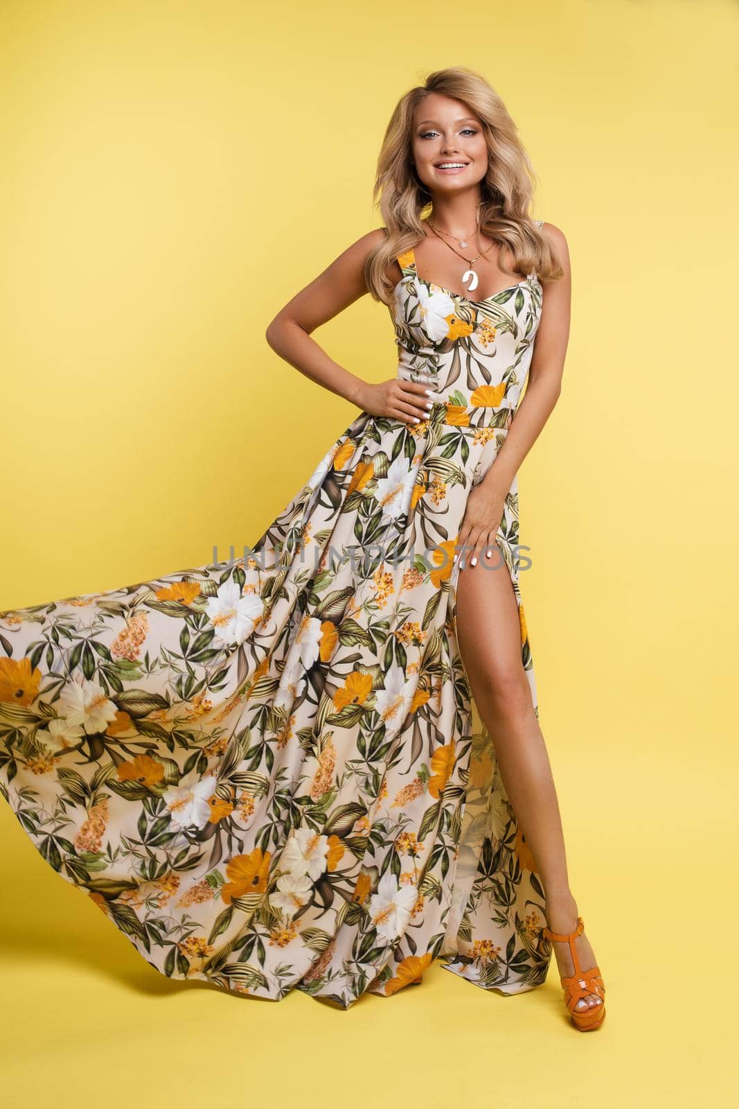 Front view of gorgeous blonde in long beautiful dress posing on yellow isolated background. Charming female with makeup and hairstyle looking at camera and smiling. Concept of beauty and summer.