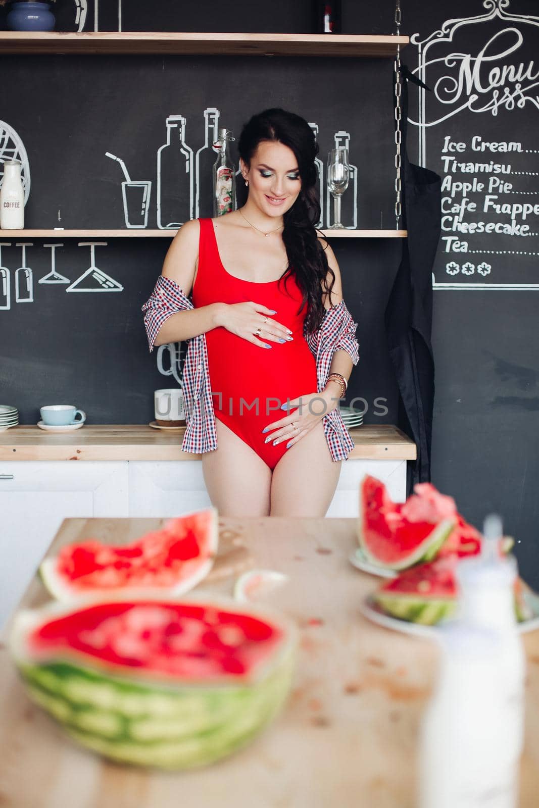 Focus on cut juicy watermelon on wooden table. Unfocused blurred background with unrecognizable pregnant brunette woman in red embracing her belly.